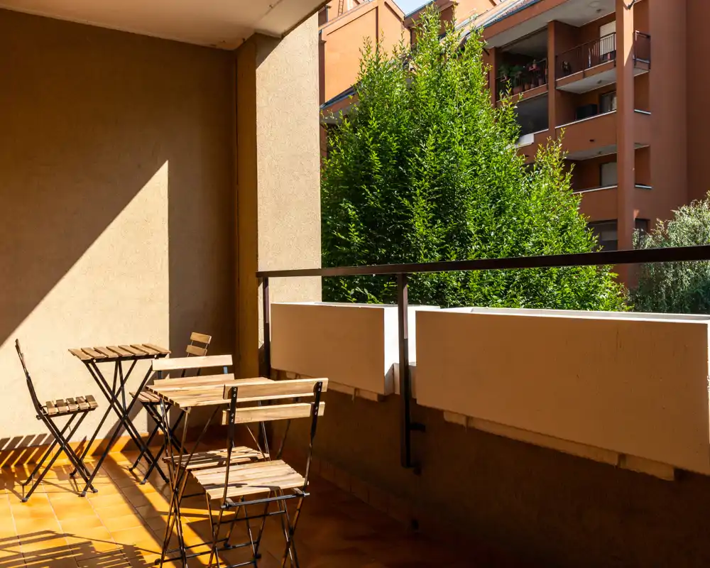 Balcón soleado con mesas y sillas bistró plegables de madera, suelo de terracota y vista arbolada — ideal para el café de la mañana.