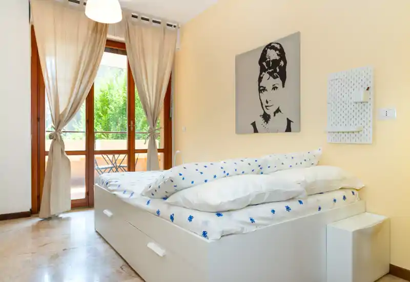 Camera da letto luminosa e ordinata con letto contenitore bianco, biancheria a fantasia e ampie porte sul balcone che illuminano la stanza con luce naturale.
