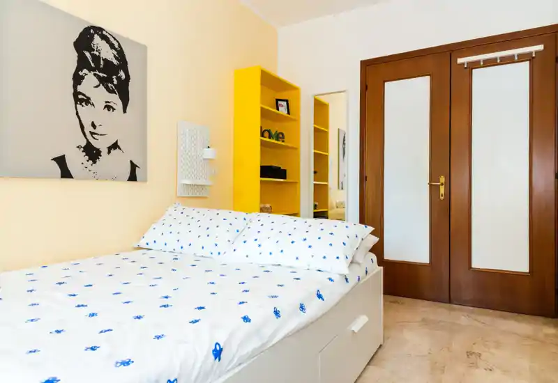Camera da letto luminosa e curata con letto matrimoniale con biancheria bianca a pois blu, scaffalatura gialla e quadro decorativo; ambiente ordinato e accogliente.
