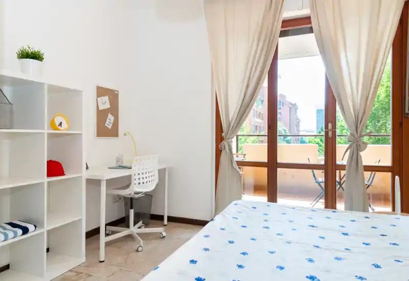 Camera da letto luminosa e ariosa con letto matrimoniale in primo piano, una scrivania e scaffalatura, e grandi porte sul balcone che lasciano entrare molta luce naturale.