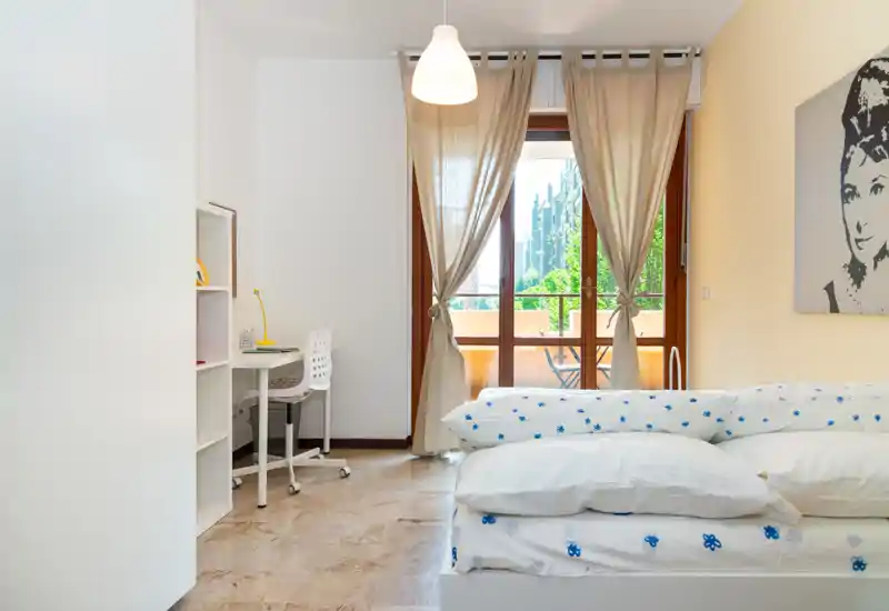 Camera da letto luminosa e ariosa con un comodo letto in primo piano, una piccola scrivania con scaffale a sinistra e un balcone con tende che filtrano la luce naturale.