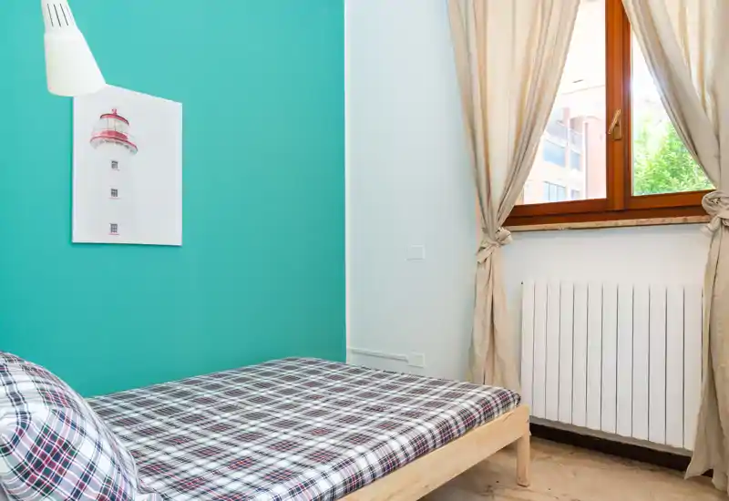 Chambre lumineuse et compacte avec un lit simple à motif contre un mur bleu sarcelle et une fenêtre apportant la lumière naturelle.