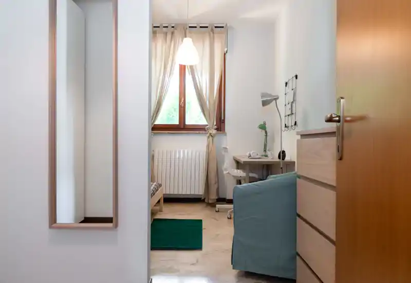 Chambre simple et chaleureuse vue depuis l'entrée, avec un petit lit, un bureau, une lampe et la lumière naturelle de la fenêtre — parfaite pour un coin travail calme.
