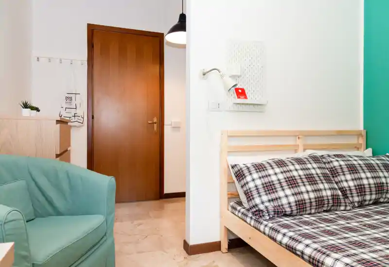 Coin chambre lumineux et sobrement décoré avec un lit simple en bois habillé d'une literie à carreaux et un fauteuil confortable à proximité ; lignes épurées et tons agréables.