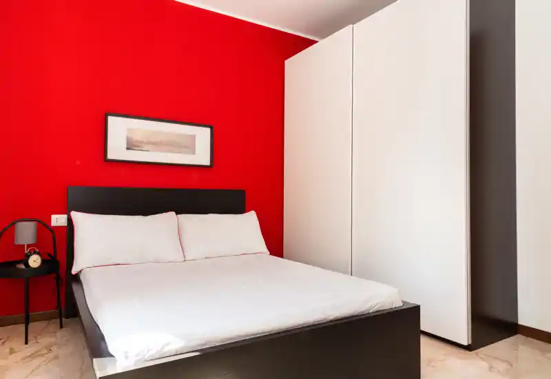 Dormitorio moderno y luminoso con cama doble frente a una pared roja intensa, cabecero negro, ropa de cama blanca y un armario blanco alto — limpio, bien iluminado y perfecto para alquileres de corta estancia.