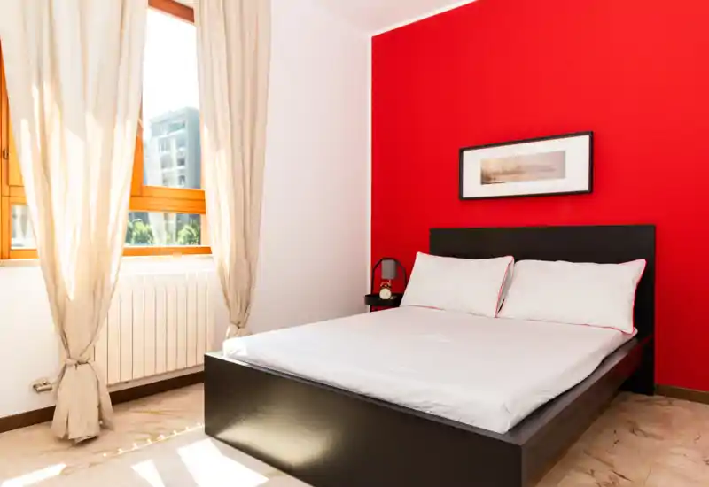 Dormitorio luminoso y moderno con cama doble, pared roja como punto focal y gran ventana que aporta mucha luz natural — ideal para anuncios de alquiler.