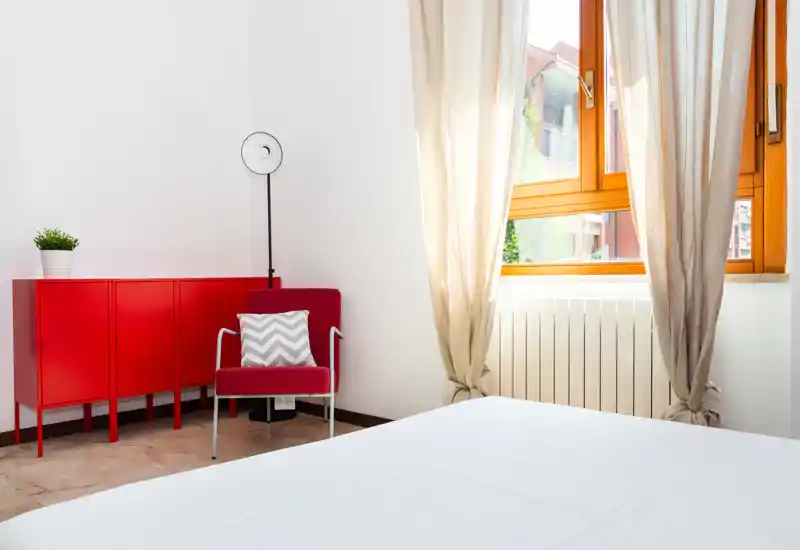 Dormitorio luminoso y minimalista con cama blanca en primer plano, gran ventana que aporta luz natural y un mueble rojo con silla a juego como acento.