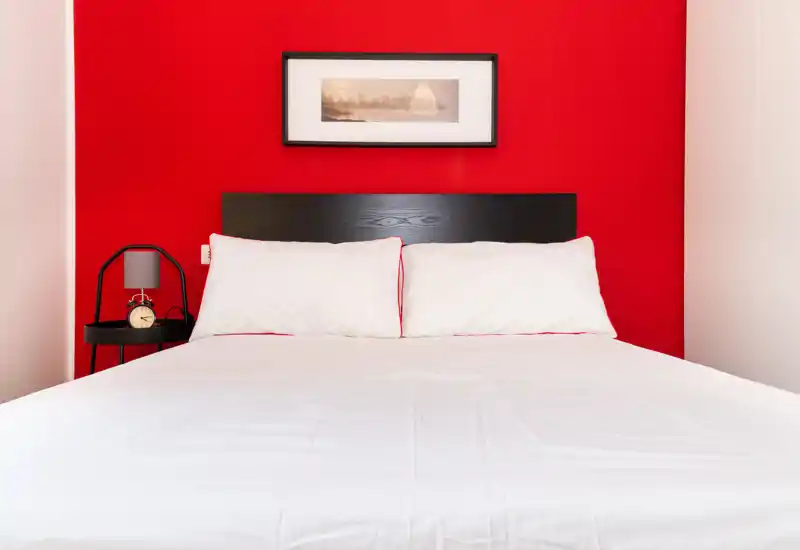 Dormitorio moderno y minimalista con una amplia cama blanca frente a una pared roja llamativa, mesita con lámpara y reloj despertador, y cuadro sobre el cabecero.