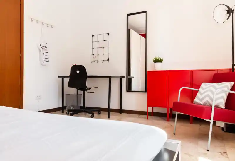 Dormitorio luminoso y moderno con cama blanca en primer plano, escritorio compacto con silla, espejo de cuerpo entero y armario rojo con un sillón a juego que añade color.