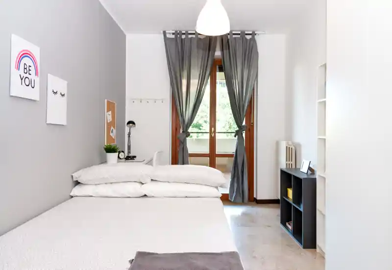 Bright, modern bedroom with a neatly made double bed, soft neutral linens, desk area and balcony doors letting in natural light — ideal for rental photos.