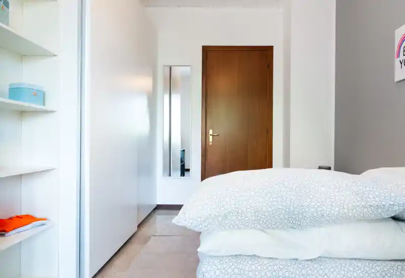 Bright, modern bedroom with a neatly made bed in the foreground, white built-in wardrobe and shelving, and a wooden door opposite; fresh linens and minimal decor create a calm, airy atmosphere.