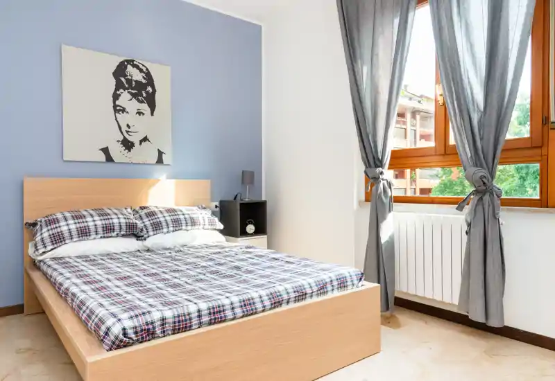 Bright, modern bedroom with a wooden double bed dressed in checked bedding, a bedside shelf with lamp and a large window letting in abundant natural light.