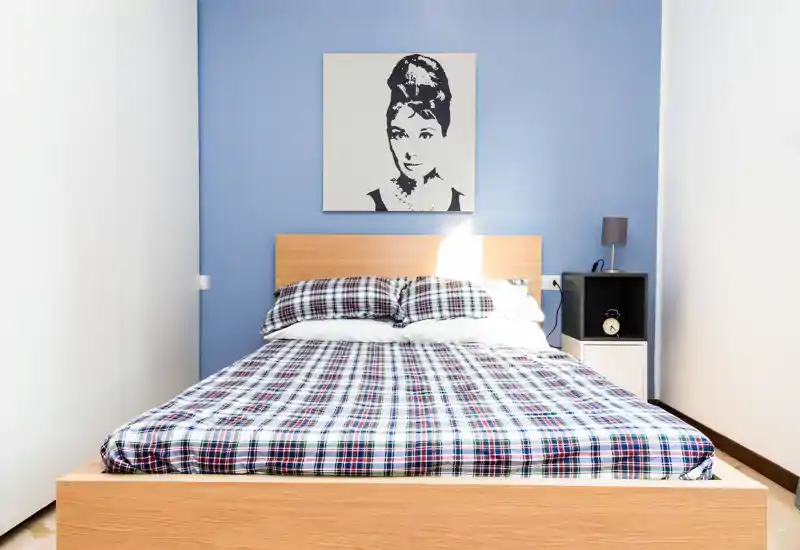 Bright, neatly made bedroom with a wooden double bed as the focal point, patterned bedding, bedside table with lamp and minimalist wall art above the headboard.