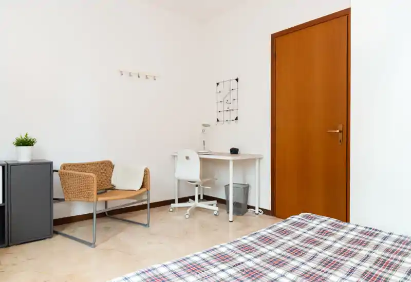 Bright, minimally styled bedroom corner with a visible bed edge, a small desk and a wicker chair — tidy and suitable for short-term rental photos.