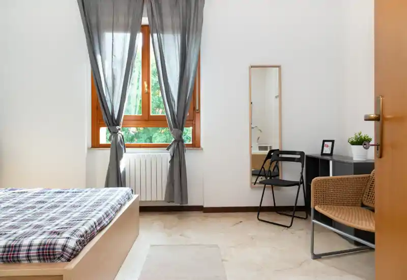 Bright, neatly staged bedroom with a wooden bed, soft plaid bedding, a window with grey curtains and a small seating/desk area — ideal for rental photos.
