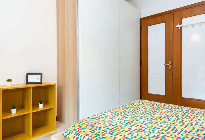 Bright, modern bedroom with a colorful duvet, built-in wardrobe and wooden door; minimal decor includes a yellow shelving unit and small plants.