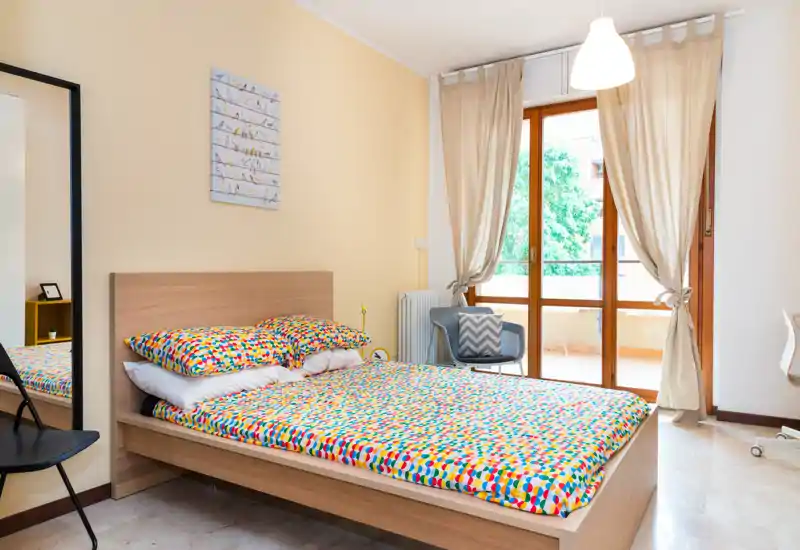 Bright, modern bedroom with a double bed, colorful bedding, a full-length mirror, balcony doors and natural light — cozy and ready for guests.