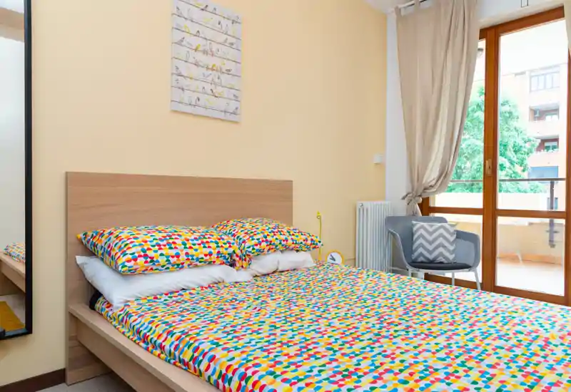 Bright, neatly styled bedroom with a full double bed dressed in colorful geometric bedding, a chair by the balcony and large window letting in natural light.