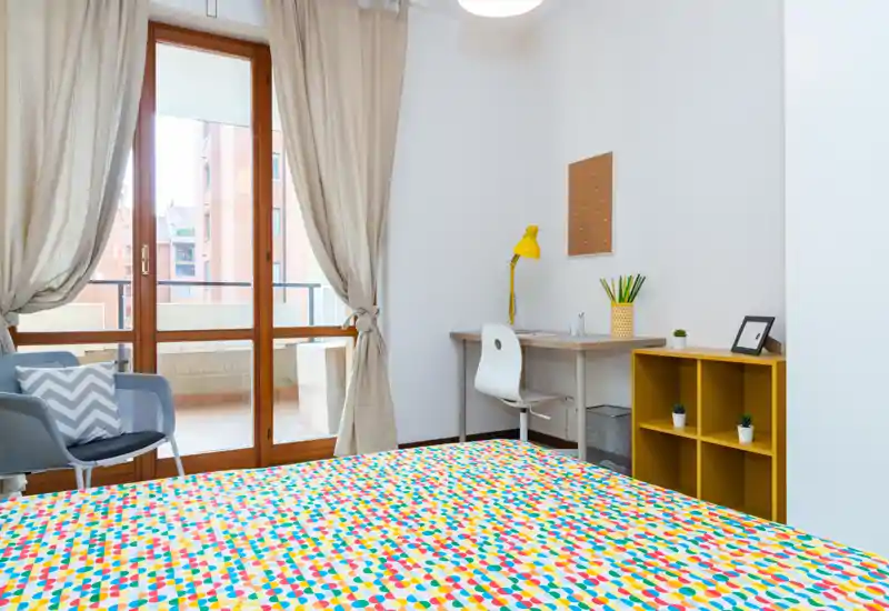 Bright, modern bedroom with a colorful bedspread, a desk and chair by the window, and a small shelving unit; large glass doors open to a balcony letting in ample natural light.