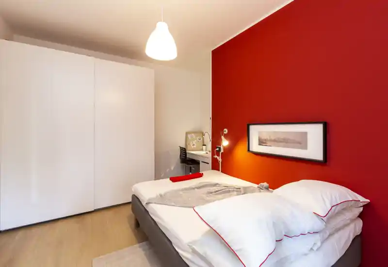 Dormitorio moderno y luminoso con pared roja de acento, cama doble con ropa de cama blanca y un pequeño espacio de trabajo en la esquina.
