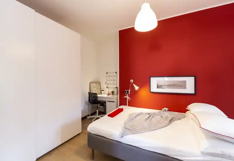 Dormitorio moderno y luminoso con cama doble frente a una pared roja, lámpara de mesita y un pequeño escritorio en la esquina — ideal para alquileres de corta estancia.