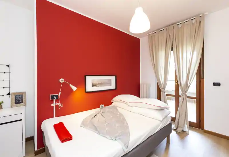 Dormitorio moderno y luminoso con pared roja como acento, cama doble con ropa blanca y textiles suaves, y luz natural desde la puerta del balcón.