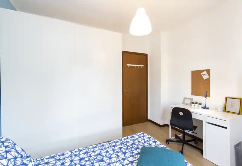 Dormitorio luminoso y moderno con colcha estampada, amplio armario blanco y pequeño escritorio — espacio limpio, funcional y bien iluminado para dormir y estudiar.