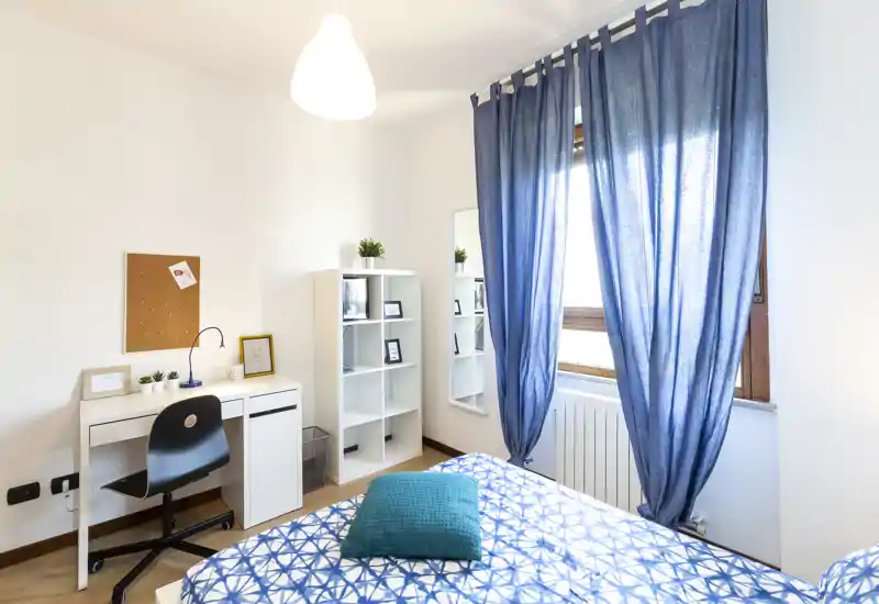 Dormitorio luminoso y bien presentado con colcha estampada, cojín turquesa y un escritorio junto a la ventana. La luz natural y las estanterías modernas crean un ambiente aireado y acogedor, perfecto para un anuncio de alquiler.