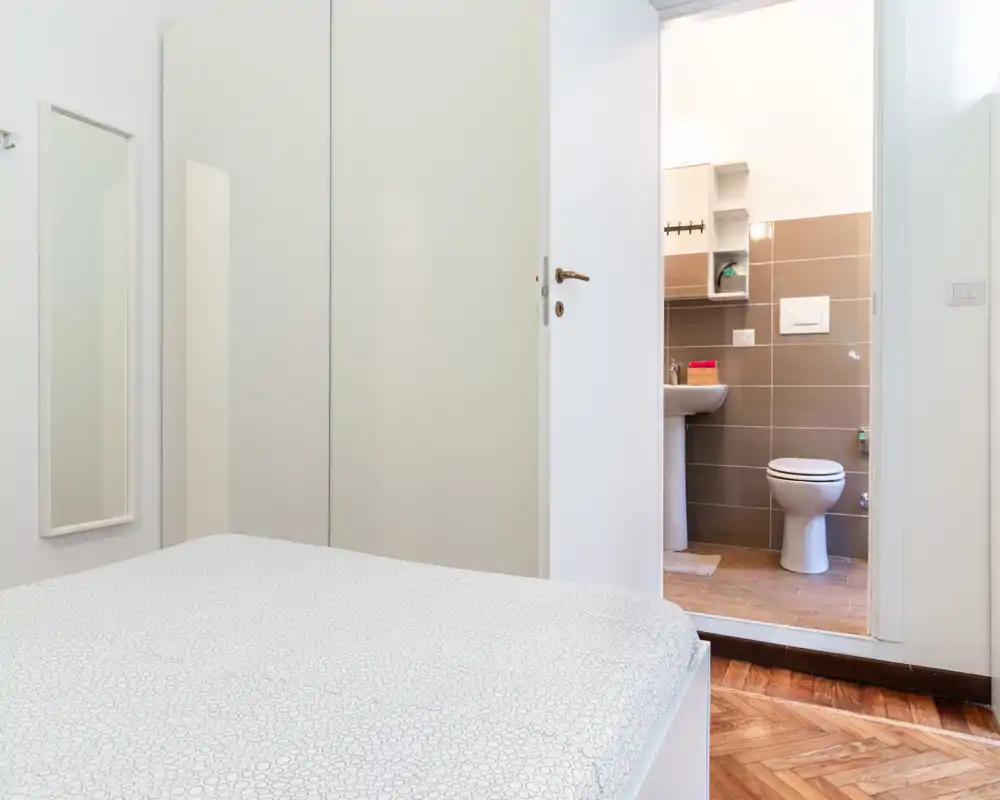 Dormitorio luminoso y minimalista que muestra la mayor parte de la cama en primer plano y un baño en suite visible por la puerta abierta. Ropa de cama neutra, suelos de parquet y armario empotrado transmiten un espacio moderno y limpio.
