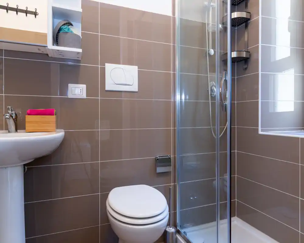 Baño moderno y compacto con lavabo de pedestal, inodoro y ducha con mampara de cristal; azulejos marrones brillantes y una ventana aportan buena luz natural.
