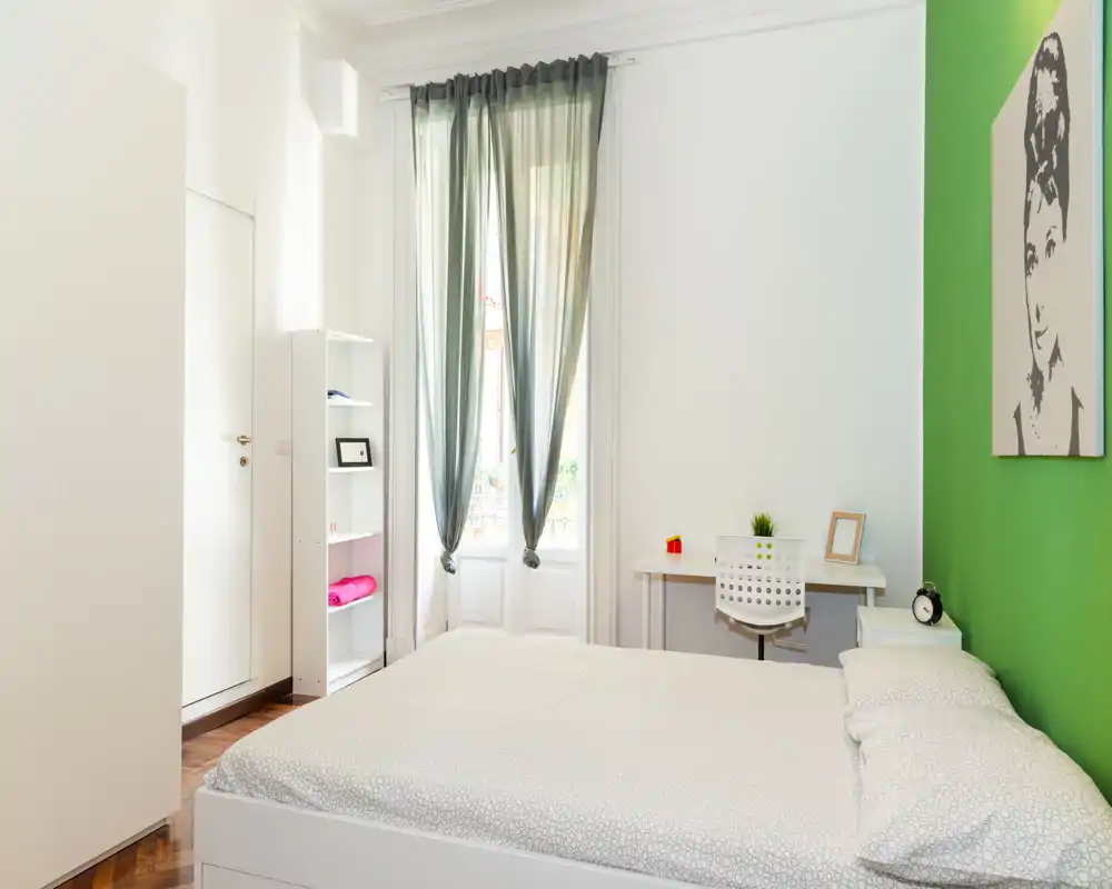 Dormitorio moderno y luminoso con cama doble, escritorio y ventana alta que aporta luz natural; pared verde que añade un toque fresco.