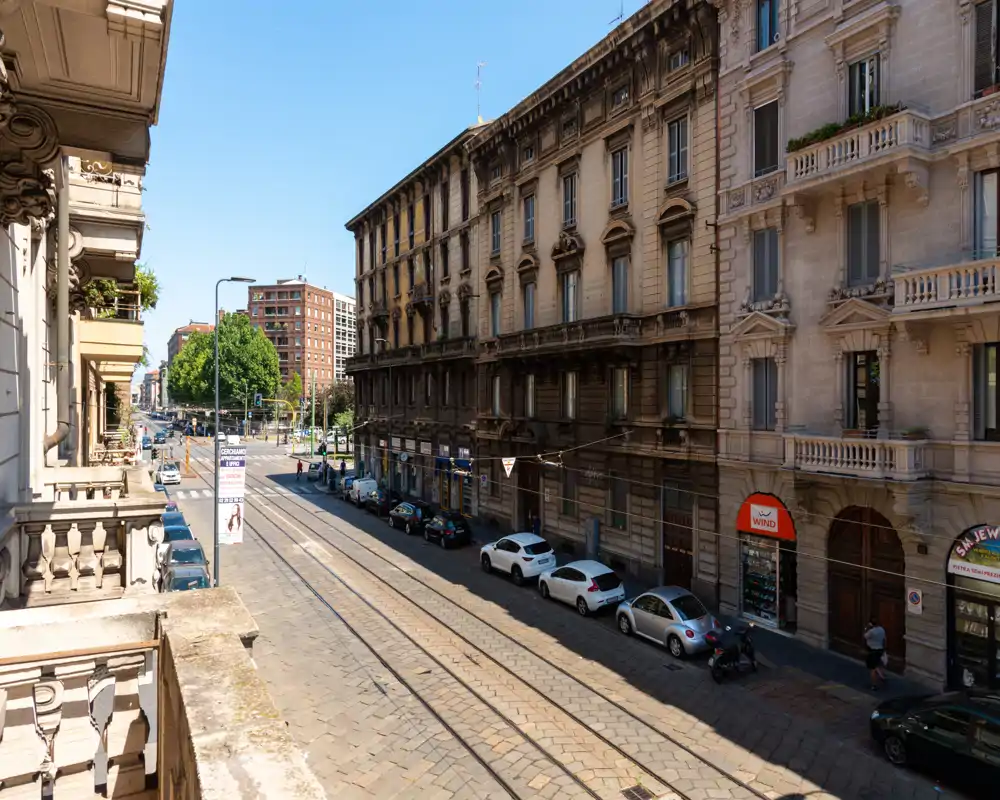 Vista soleada desde un balcón con las vías del tranvía, fachadas históricas y coches aparcados — ideal para mostrar el barrio y las conexiones.