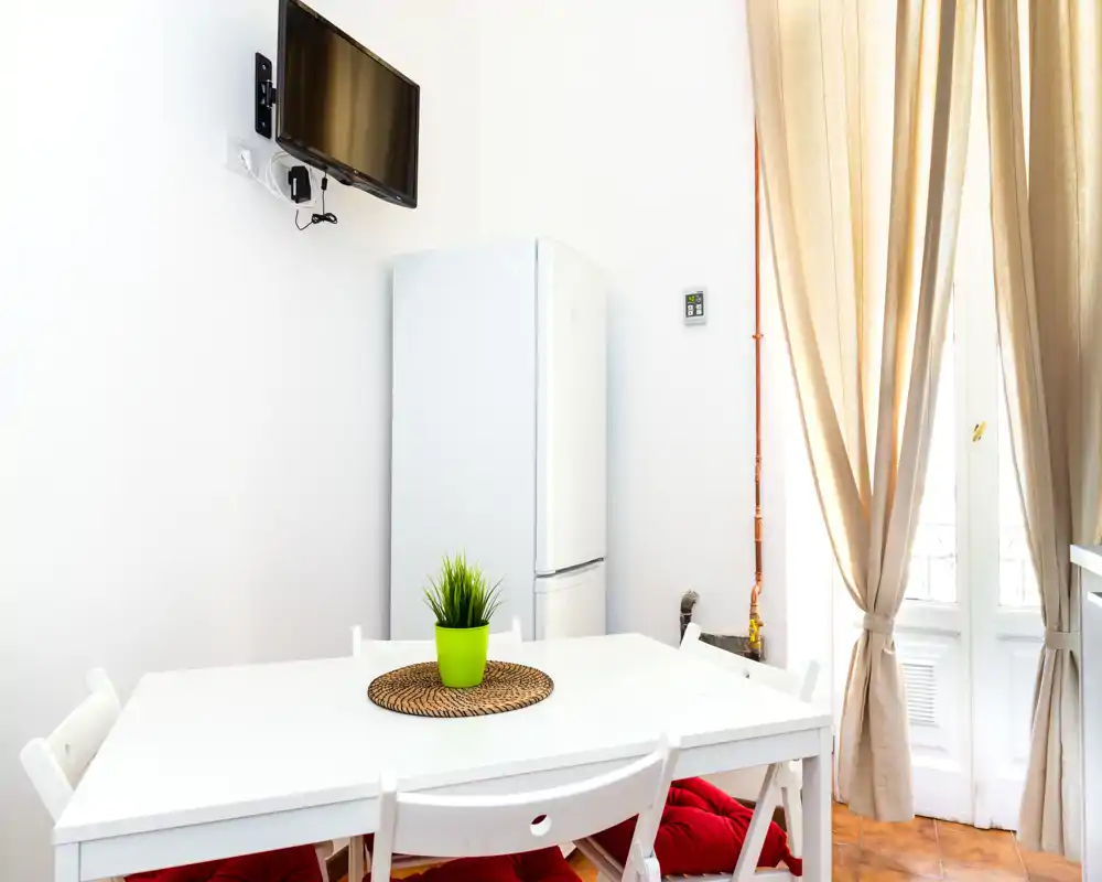 Cocina compacta y luminosa con mesa blanca, frigorífico y luz natural desde la puerta al balcón; decorada con una planta pequeña y un salvamanteles.