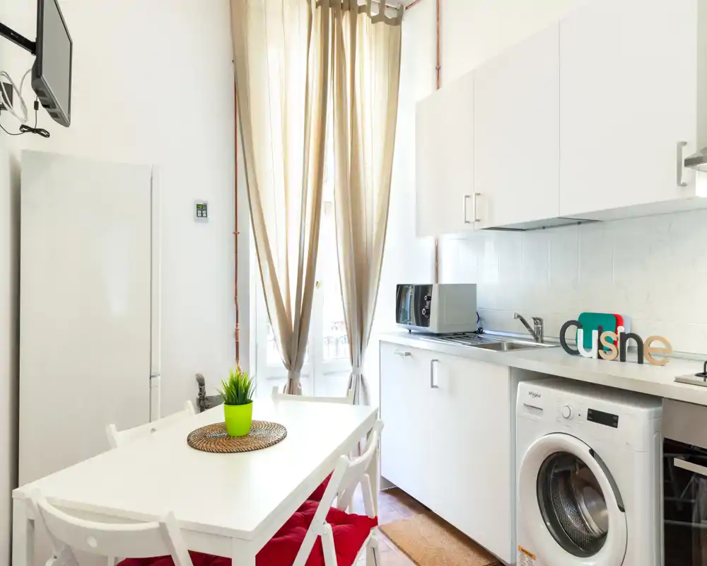 Cocina compacta y luminosa con muebles blancos, mesa comedor, lavadora y luz natural desde la ventana alta — perfecta para un apartamento pequeño.