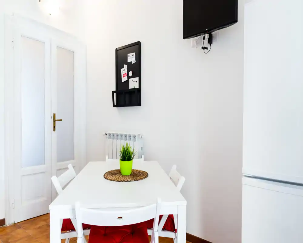 Zona de comedor luminosa y minimalista con mesa blanca, cuatro sillas con cojines rojos, una pequeña planta decorativa y TV en la pared — ideal para comidas informales en un espacio compacto.
