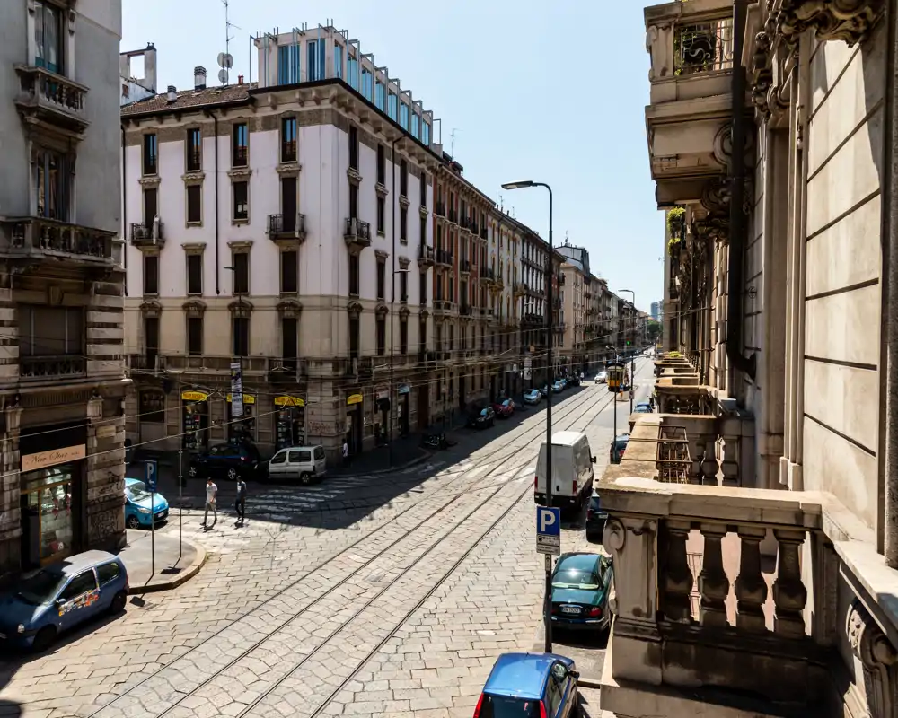 Balcón soleado con vistas a una calle empedrada con vías de tranvía y fachadas clásicas de Milán — ideal para mostrar la ubicación y el encanto urbano.