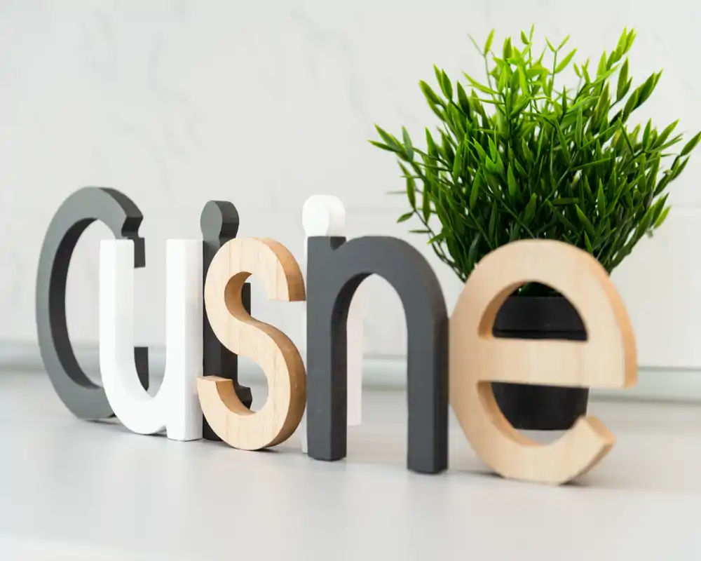 Plano cercano de letras decorativas de madera que forman 'Cuisine' con una pequeña planta en maceta: un detalle brillante y cuidado que aporta calidez a la cocina.