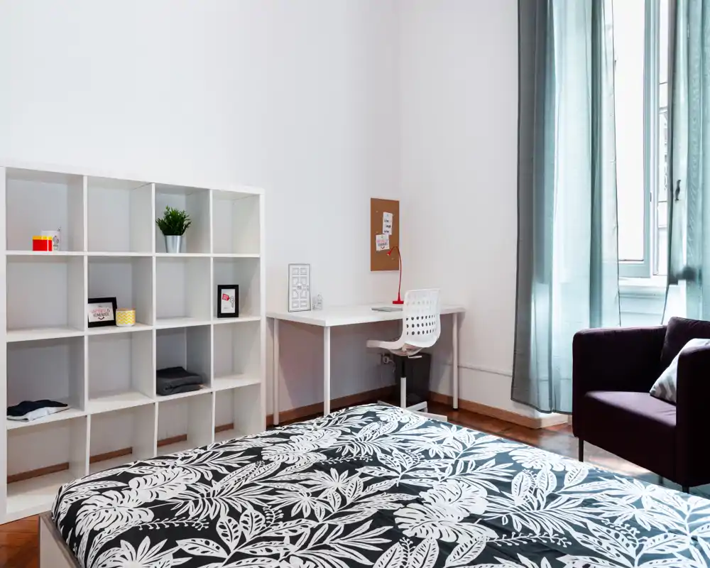 Camera da letto moderna e luminosa con copriletto nero e bianco a motivo, scrivania e sedia per lavorare, scaffalatura per riporre oggetti e una poltrona accogliente vicino alla finestra.