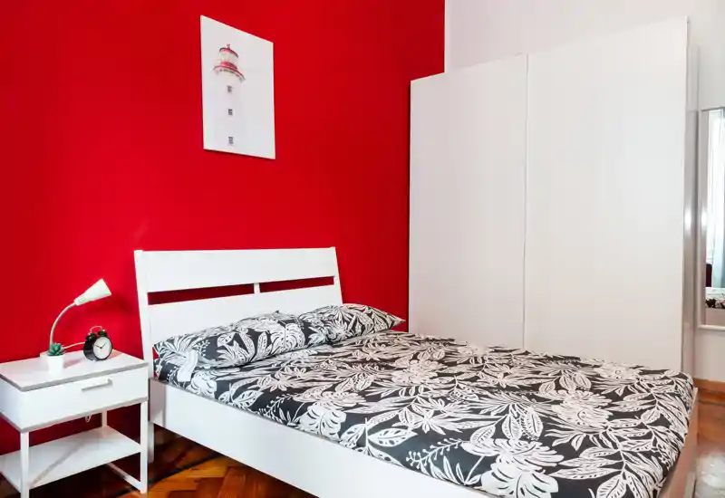 Dormitorio moderno y luminoso con estructura de cama blanca y pared roja de acento. La ropa de cama en blanco y negro con estampado, la mesita con lámpara y el amplio armario blanco crean un aspecto limpio y contemporáneo, perfecto para anuncios.
