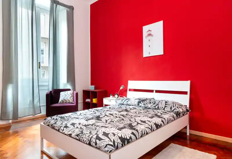 Dormitorio moderno y luminoso con estructura de cama blanca, ropa de cama estampada y una pared roja llamativa. Luz natural de una ventana alta, sillón acogedor y mesita de noche que hacen el espacio ideal para el anuncio.