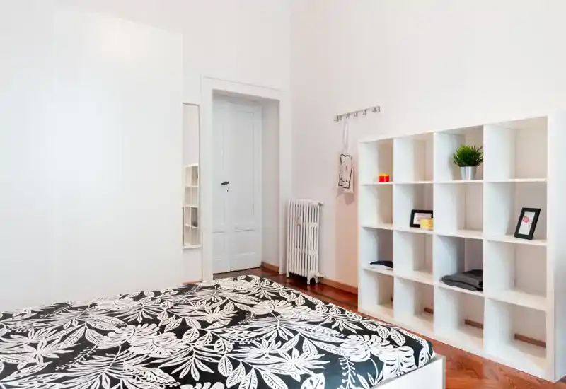 Dormitorio luminoso y minimalista con colcha estampada, estantería blanca y armario — líneas limpias y suelo de madera cálida que crean un ambiente moderno y sereno.