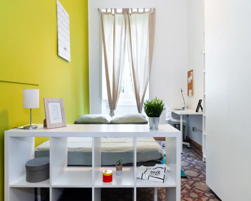 Chambre moderne et lumineuse avec un lit double, une étagère au pied du lit et un coin bureau près de la fenêtre. La lumière naturelle et le mur vert apportent une ambiance fraîche et accueillante.