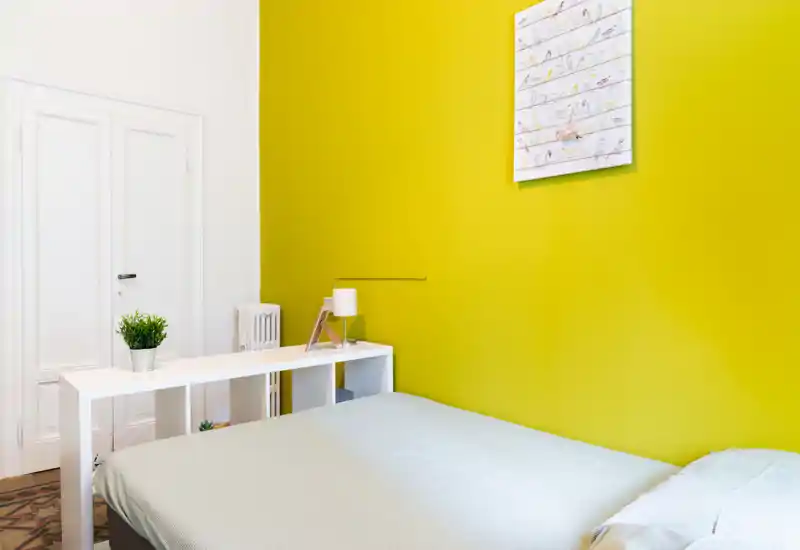 Camera da letto luminosa e moderna con un grande letto contro una vivace parete gialla, mensole minimal dietro il letto e una semplice lampada da comodino. Linee pulite e luce naturale rendono la stanza ariosa e accogliente.