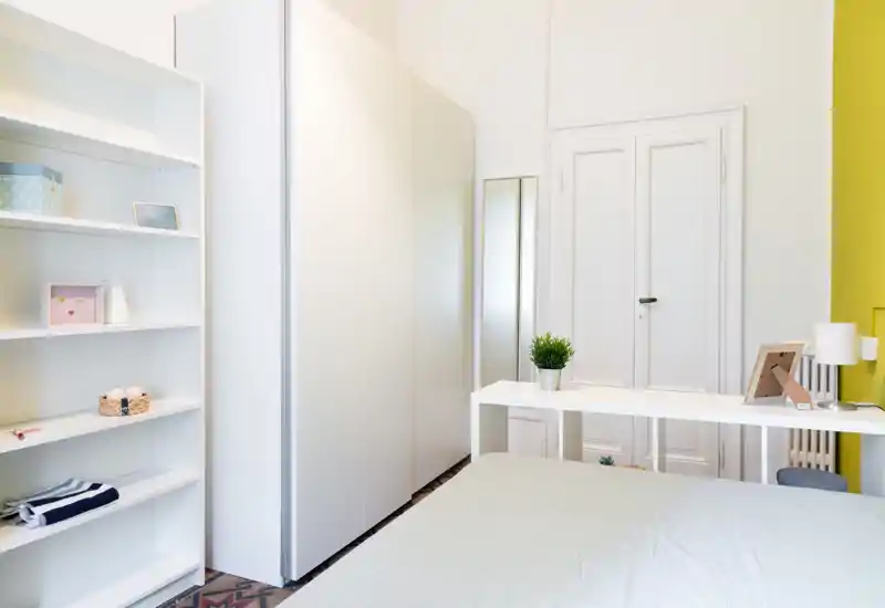 Camera da letto luminosa e minimal con letto visibile, grande armadio e una piccola scrivania. Arredi bianchi e puliti, una pianta decorativa e luce naturale soffusa rendono l’ambiente accogliente per gli annunci.