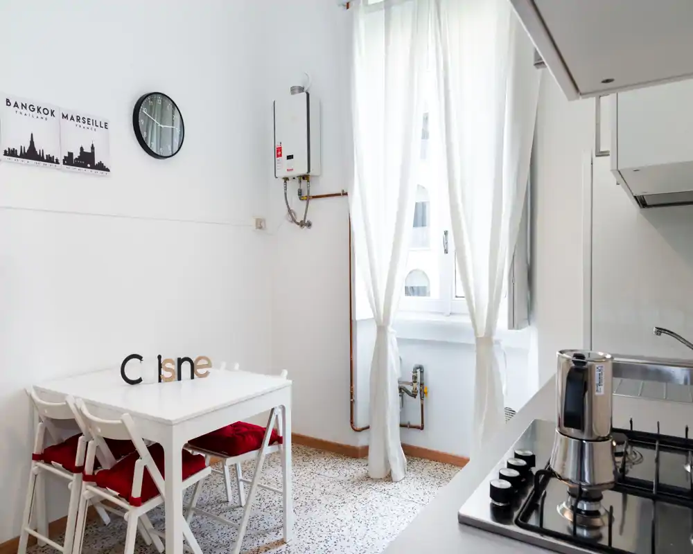 Cucina compatta e luminosa con piccolo tavolo da pranzo, quattro sedie con cuscini rossi, piano cottura e finestra che illumina l'ambiente: pulita e minimal.