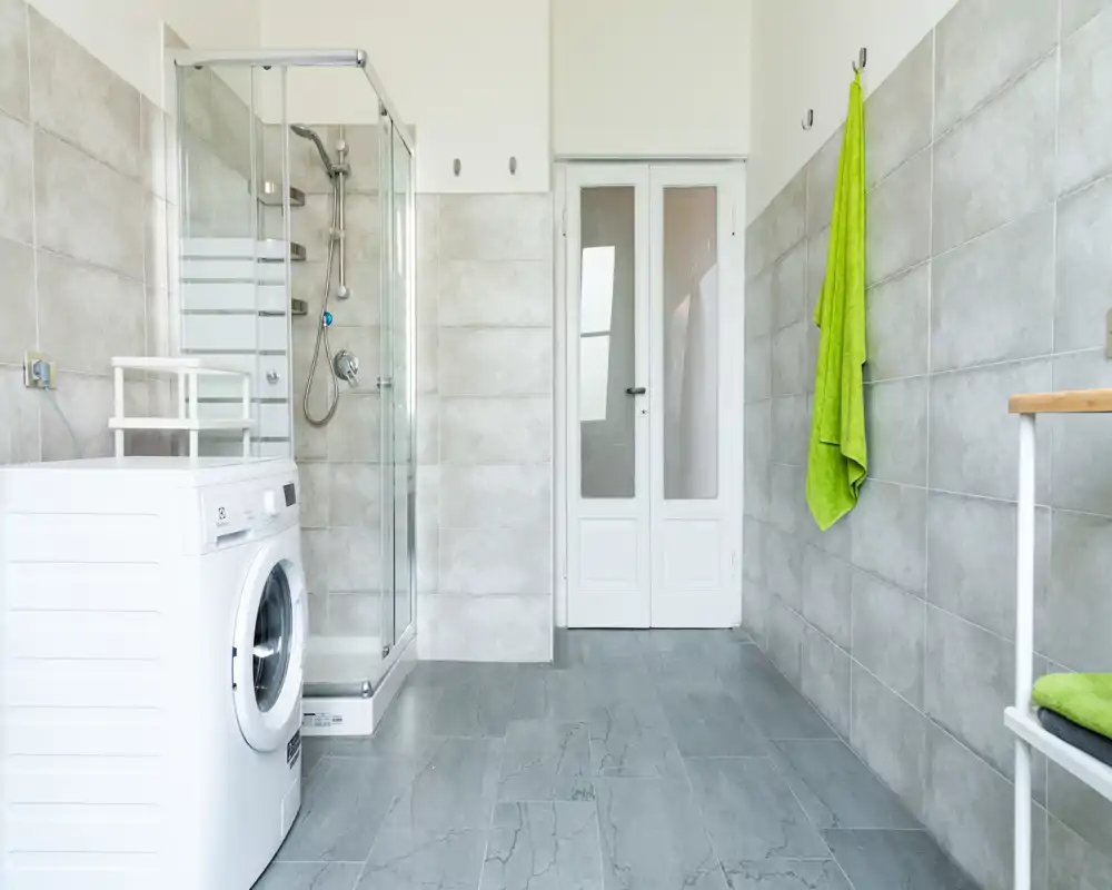 Bagno luminoso e moderno con pareti piastrellate, cabina doccia in vetro e lavatrice — ambiente pulito e funzionale.