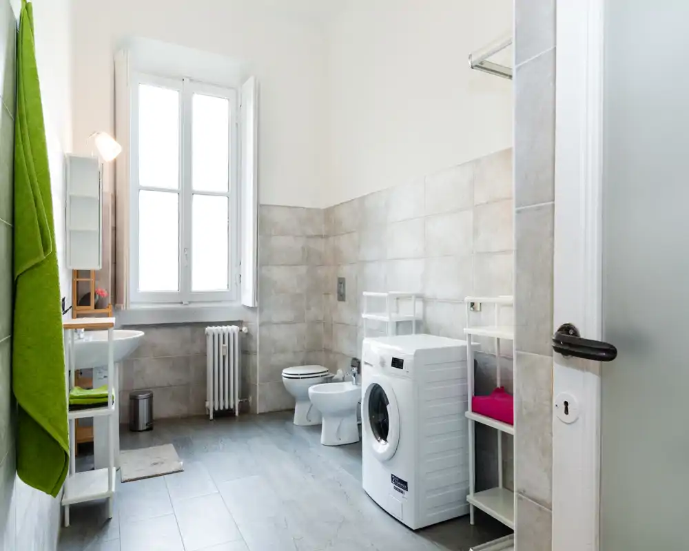 Bagno moderno e luminoso con ampia finestra, lavatrice, bidet e WC; piastrelle grigie chiare e scaffalature minimali per un ambiente funzionale e ordinato.