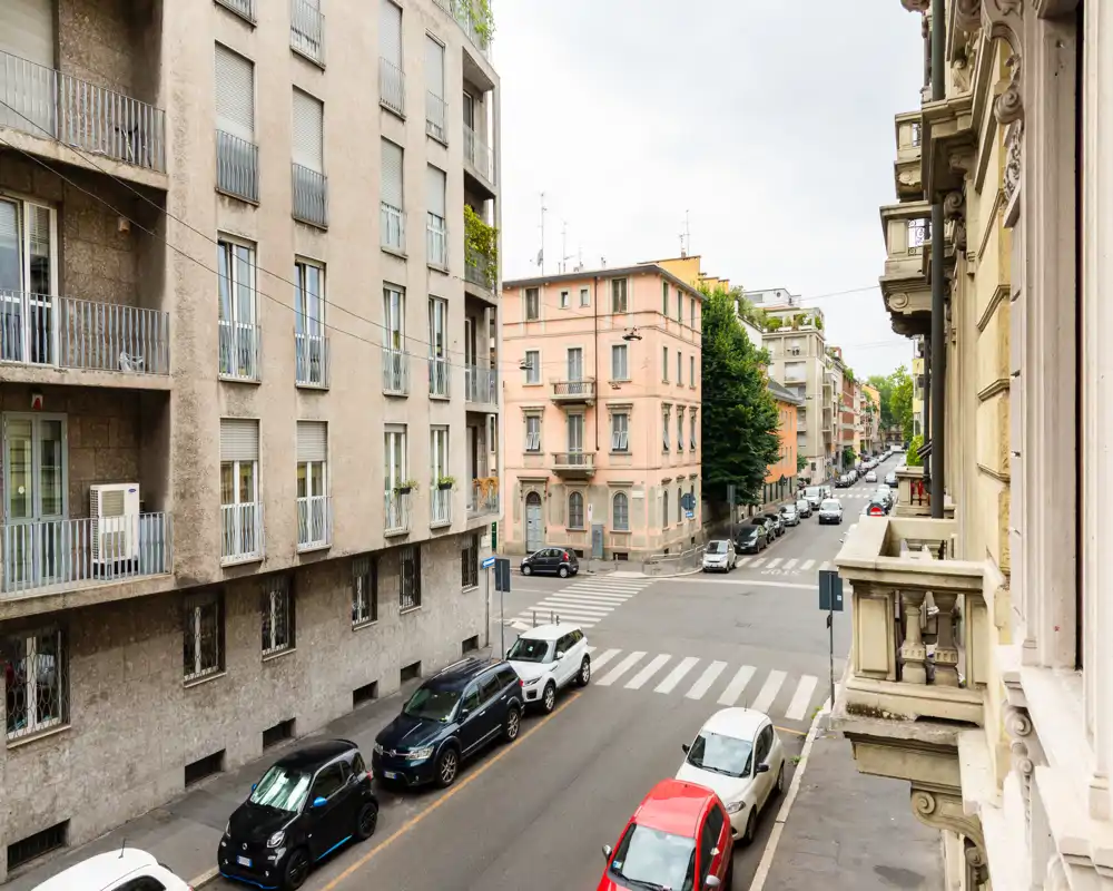 Vista sulla strada cittadina da un balcone con auto parcheggiate, attraversamenti pedonali e palazzi circostanti — ideale per pubblicizzare un appartamento centrale con affaccio sulla strada.
