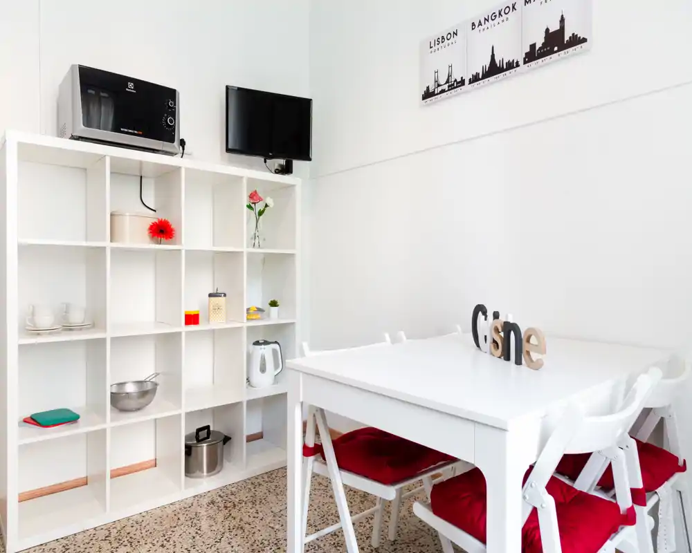Zona pranzo luminosa e compatta con tavolo bianco e quattro sedie con cuscini rossi, scaffalatura aperta con accessori da cucina e piccoli elementi decorativi.