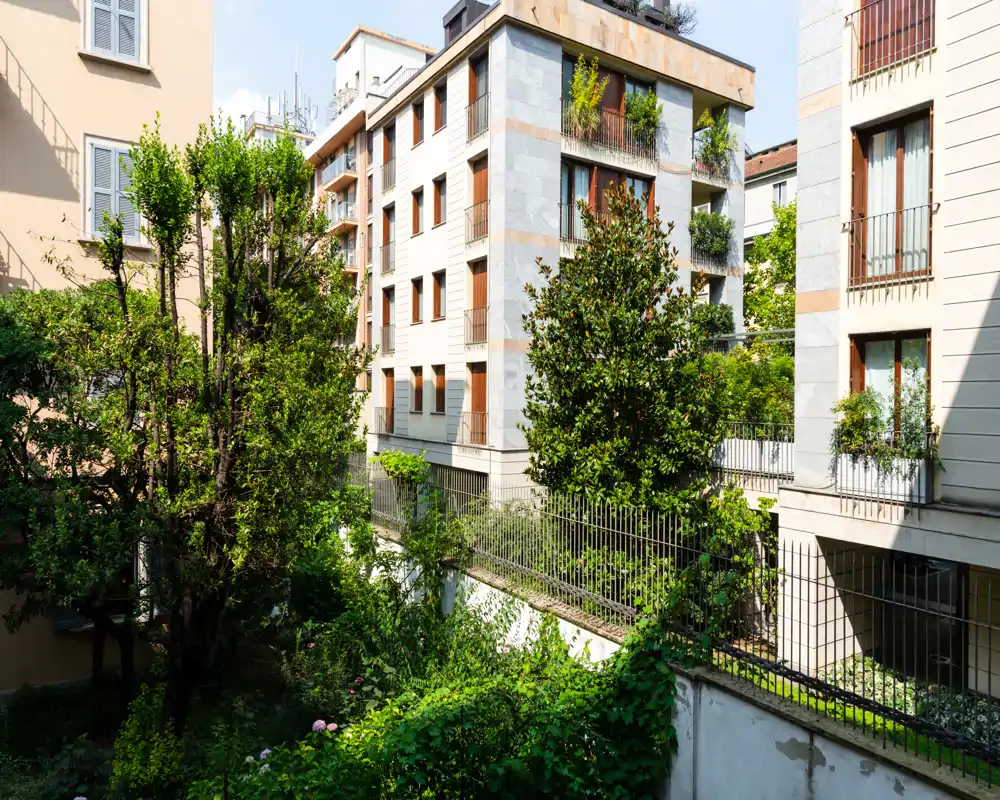 Vista sul cortile soleggiato con alberi maturi e verde paesaggistico di fronte a edifici residenziali — ideale per descrivere giardini comuni o l’affaccio dell’appartamento.